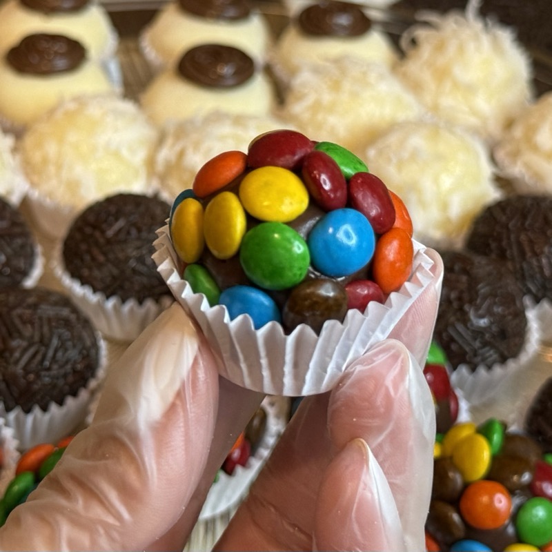 Hand holding a brigadeiro covered in colorful candy pieces above a tray of assorted flavors