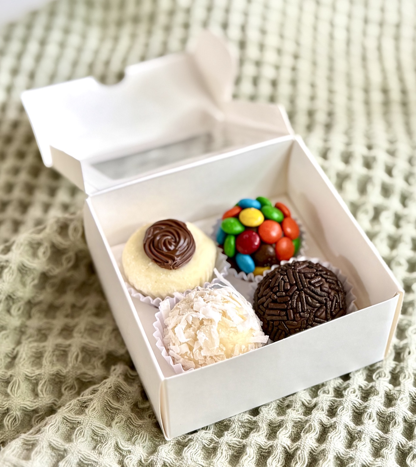 Open gift box of four assorted brigadeiros on a sage waffle blanket